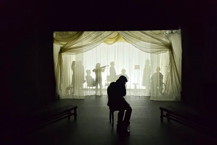Il Monumento di Eduardo De Filippo, foto di scena: un uomo seduto in controluce davanti a un sipario di tulle con figure in ombra